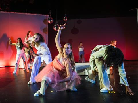 TRAUER//FALL, POLYLUX, Schauspiel Köln – Auf dem Bühnenfoto sind in etwas sieben Personen abgebildet. Die rechte seite des Bildes schimmert in einem grünen Licht und die linke in einem roten. Im Vordergrund ist eine weiblich gelesene Person die ein rosa Kleid trägt und hockt. Sie schaut nach vorne aber nicht in die Kamera, sie hat ihren Arm elegant über sich ausgestreckt. Links und rechts von ihr aber weiter hinten sind noch zwei Personen die ebenfalls in interressanten Positionen verharrt sind. Bis nach ganz hiinten der Bühne geht das weiter so.