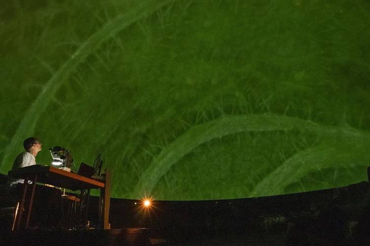 »Unsichtbare Welten: Vom Mikrokosmos zum Universum«: Mega-Mikroskopie im Planetariumssaal des Zeiss-Großplanetariums. – Mann mit einem Mikroskop sitzt auf der Bühne, im HIntergrund die große Kuppel eines PLanetariumssaals.