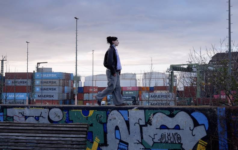 © Julia Krewani und Ralph Etter – Eine Person läuft in beginnender Dunkelheit auf einer Mauerkante, im Hintergrund Container.