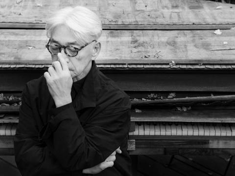 Ryuichi Sakamoto, Portrait – Schwarzweiß-Fotografie eines nachdenklichen Menschen mit Brille und hellem Haar vor einem alten, teils kaputten Klavier mit Herbstblättern.