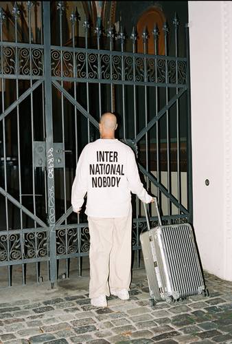 Foto: Lee Everett Thieler – Eine Person mit Glatze, in hellem Outfit, stehen vor einem verschlossenen Tor mit Koffer. Auf dem Ruecken steht "INTERNATIONAL NOBODY".