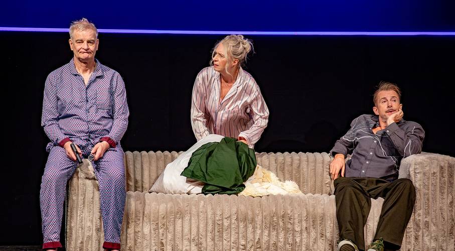 Was ist los mit der Welt?! – Drei Personen in Schlafanzügen auf einem Sofa, die linke Person hält ein Smartphone, die mittlere hat hellgraues Haar, die rechte Person stützt den Kopf mit