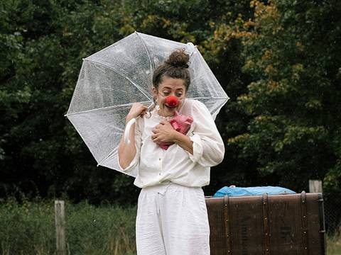  – Person mit roter Clownsnase hält durchsichtigen Regenschirm und roten Stoff, trägt weiße Kleidung. Hintergrund aus Bäumen und einer Holzkiste.