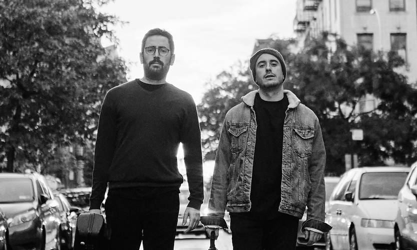 Alejandro Oliván & Carlos Gil-Gonzalo – Zwei Personen gehen nebeneinander eine Straße entlang. Eine trägt einen Pullover, die andere eine Jeansjacke und Mütze. Autos parken am Straßenrand.