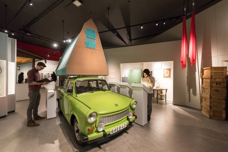 Dauerausstellung "Alltag in der DDR" – ein grüner Trabant mit aufgebautem Dachzelt steht in der Ausstellung "Alltag in der DDR", an den Seiten stehen Besucher und lesen die Informationen zum Exponat