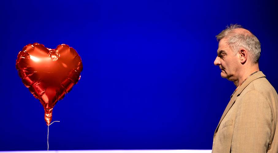 Was ist los mit der Welt?! – Roter Herzballon schwebt links. Person im Profil nach rechts blickend, mit beigem Sakko. Blauer Hintergrund.