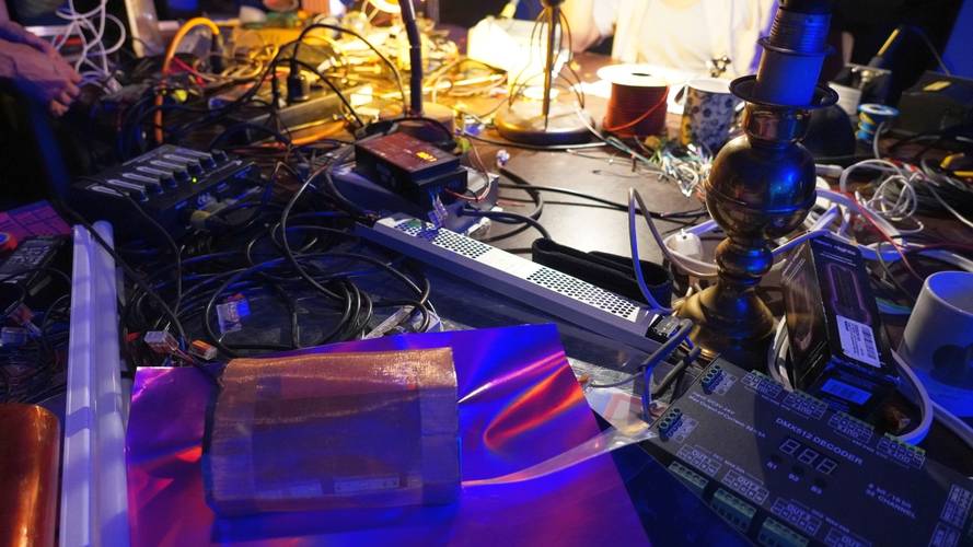 Ein Tisch voller Materialien - Kabel, reflektierende Folien, unterschiedliche Haushaltslampen, Drähte, oben links ein paar Hände.