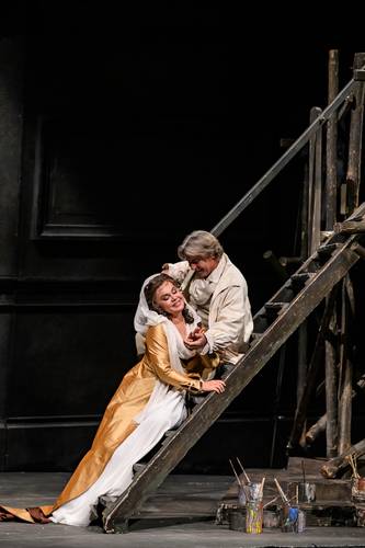 Tosca – Männlich gelesene Person auf Holzgerüst hält weiblich gelesene Person in Gold- und Weißkleid in den Armen; Bühne mit Malutensilien.