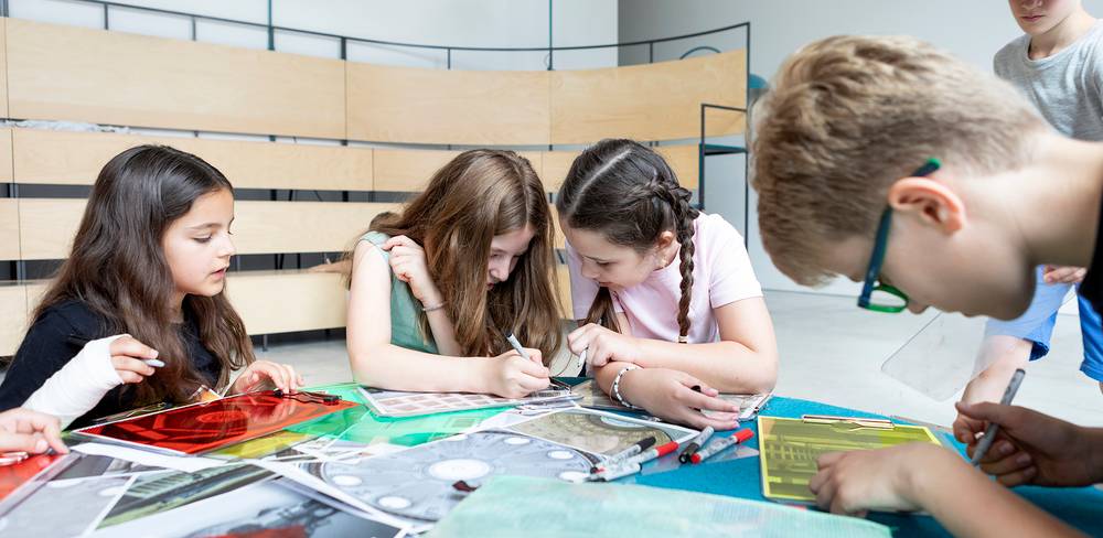 Fünf Kinder sitzen an einem Tisch und sind in eine kreative Aktivität vertieft, mit Papieren, Stiften und durchsichtigen Farbfolien.