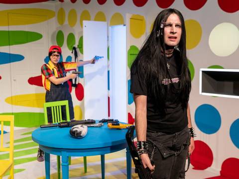  – Zwei Personen in einem Raum mit bunten Punkten. Eine Person im Hintergrund mit Mütze und Spielzeugpistole. Tisch mit Waffen und Schädel.