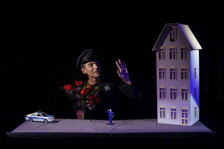 Blue – Person in Uniform mit Blumenstrauß, Modellauto, blau-weiße Figur und Modellhaus auf Tisch vor schwarzem Hintergrund.