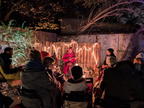  – Gruppe von Menschen sitzt im Halbkreis um Lagerfeuer im Garten, dahinter leuchten Lichter am Zaun.	person in roten Kleidern spricht.