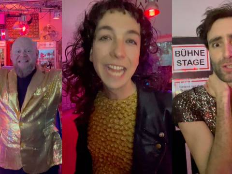  – Drei Personen in bunter Kleidung. Zwischen ihnen ein Schild mit "Bühne Stage". Rote Lampen im Hintergrund.