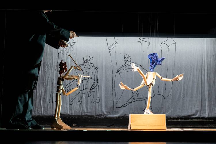 Die Geschichte vom Soldaten, Salzburger Marionettentheater – Zwei Holzkasper stehen auf einer Bühne, ein Mensch bedient die Fäden, Hintergrund Leinwand mit Zeichnungen.