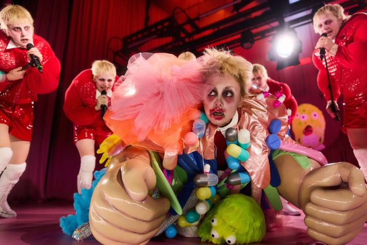 Die Welt im Rücken – Bühnenperformance mit mehreren Personen in glitzernden roten Kostümen; im Vordergrund eine farbenfroh verkleidete Figur mit großen, überdimensionierten Requisitenhänden und buntem Dekor, umgeben von grellem Bühnenlicht.