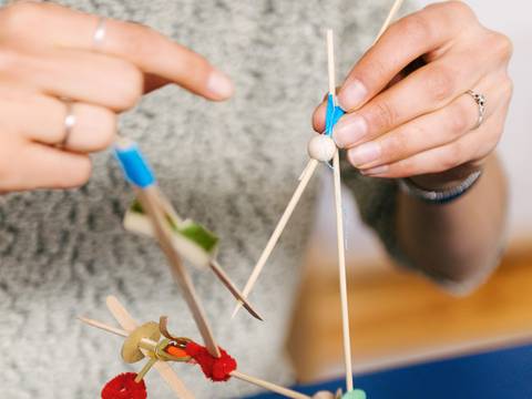 – Foto: Nahaufnahme von zwei Händen, die eine Art Mobile aus verschiedenen Materialien wie Kork, Holz und Papier in halten.