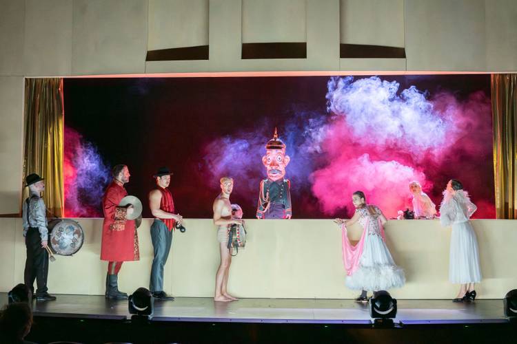 Der Hauptmann von Köpenick – Männer und Frauen mit Instrumenten stehen vor einer Art Kasperletheater mit einer uniformierten Figur mit Pappmachékopf