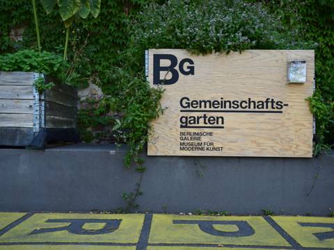 Gemeinschaftsgarten auf dem Gelände der Berlinischen Galerie – Foto: Ein Holz-Schild trägt die Aufschrift „Gemeinschaftsgarten“ und das Logo der Berlinischen Galerie. Um das Schild herum wachsen Pflanzen. Links daneben ist ein weiterer Pflanzkübel zu sehen.