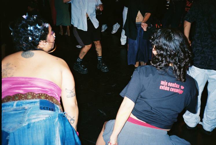Fotos: Inmensidades – Personen tanzen in einem Club. Eine Person trägt ein T-Shirt mit der Aufschrift "mis santxs estan convmxg".
