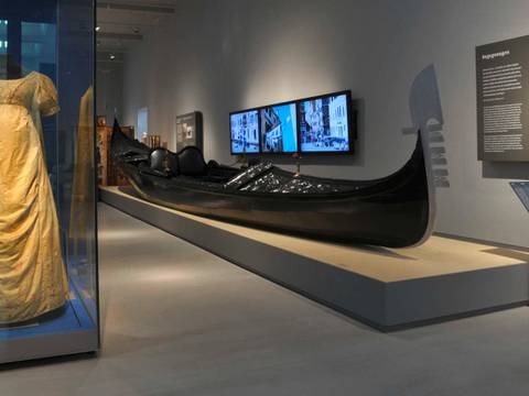 Ansicht der Dauerausstellung „Kulturkontakte. Leben in Europa“ – Eine Ausstellungsraum mit einer gelben Kleidvitrine und einer schwarzen Gondel. Rechts Texttafel: "Begegnungen", "Encounters". Wandbildschirm zeigt Kanäle.