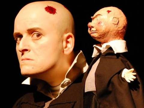  – Eine Person mit Glatze und falscher Wunde trägt eine Puppe auf der Schulter. Die Puppe hat eine ähnliche Glatze und Kleidung.