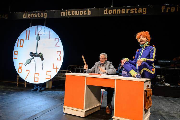 Buehne mit grosser Uhr, Mann am Schreibtisch, Person im bunten Kostuem sitzt darauf. Wochentage: dienstag, mittwoch, donnerstag.