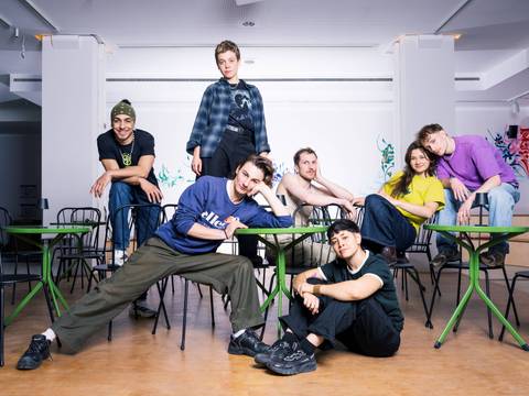  – Sieben Personen posieren lässig auf und um grüne Tische und schwarze Stühle, bunte Wandmalerei im Hintergrund. Eine Person trägt ein Shirt mit "ellesse".