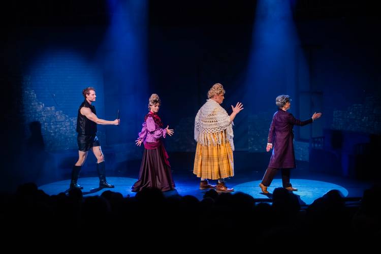Dracula – A Comedy of Terrors – Vier Personen in Kostümen in Reihe auf einer Bühne mit blauem Licht. Links trägt eine Person schwarze Kleidung, rechts lila und gelb karierte Kleidung.