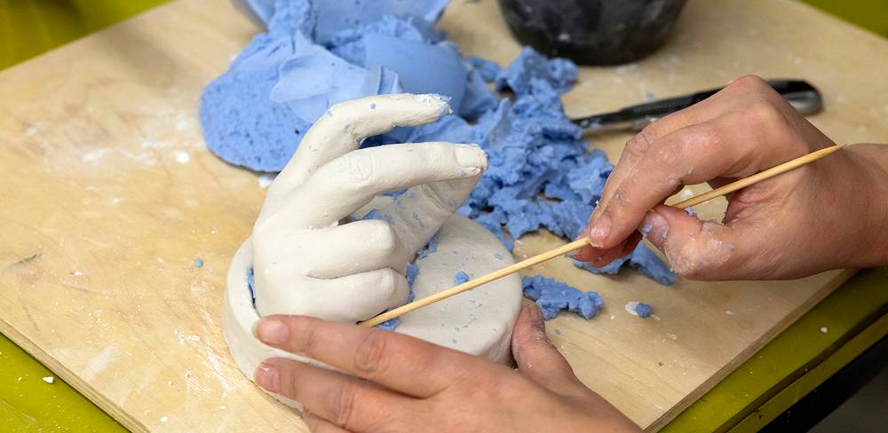 Zwei Haende bearbeiten eine weiße Gips-Handform auf Holzplatte, umgeben von blauen Silikonbruchstuecken und einem Werkzeug.