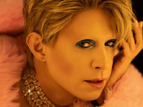 – Androgyn wirkende Person mit blonden Haaren, starkem Make-up, rosa Kunstfell und funkelndem, steinbesetztem Halsband schaut direkt in die Kamera.
