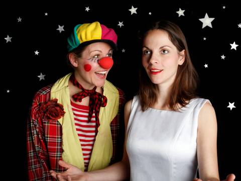  – Zwei Personen vor schwarzem Hintergrund mit Sternen. Links Clown mit rotem Hut, roter Nase und kariertem Mantel. Rechts Person im weissen Kleid, neutraler