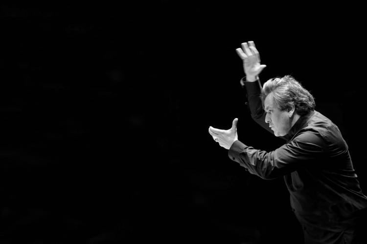 Sir Antonio Pappano, Chefdirigent des London Symphony Orchestra – Am rechten Bildrand ist vor schwarzem Hintergrund ein Mann zu sehen, der beim Dirigieren die Arme vor sich in die Höhe hält.