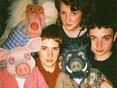  – Drei Personen und drei Handpuppen in bunter Kleidung, zwei Bierkrüge. Eine Person hinter den Puppen, eine links, eine rechts, alle schauen in die Kamera.