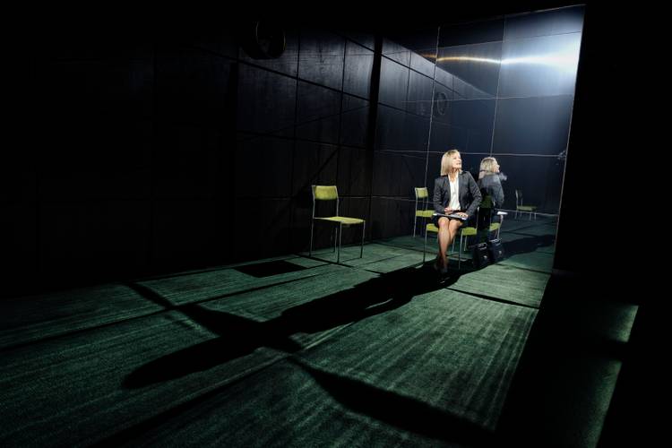 Glücklich die Glücklichen – Person in Anzug auf grünem Teppich sitzt vor Spiegelwand, bewirkt starke Licht- und Schattenspiele; zwei leere Stühle im Hintergrund.