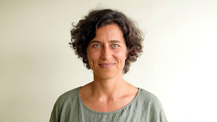 Theresa Adam – Lächelndes Gesicht einer Frau mit lockigem Haar, die ein graues Oberteil trägt, vor hellem Hintergrund.