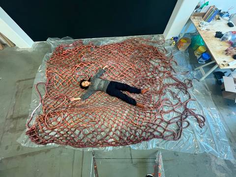 Jiyoung Yoon, There was a time when, not knowing how to live, I took out my entrails to make a net (2025). Courtesy the artist – Person liegt auf einem großen, roten Netz aus Seilen, das auf einer durchsichtigen Plane ausgebreitet ist. Links Tisch mit Werkzeugen und Materialien.