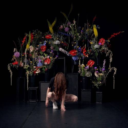 THE PRESSING 2 – Eine Person kniet mit gesenktem Kopf vor einem aufwändigen, bunten Blumenarrangement.
