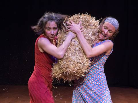  – Zwei person kämpfen um einen Strohbündel, eine in rotem Kleid, die andere in blauem Kleid mit Kopftuch auf dunklem Hintergrund.
