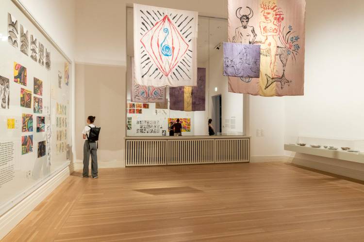 Gabriele Stötzer: "Dabei sein und nicht schweigen", Installationsansicht, Gropius Bau, 2026 – Besucher betrachtet moderne Kunstwerke von der Künstlerin Gabriele Stötzer in einer Galerie mit Holzfußboden. An den Wänden hängen abstrakte Zeichnungen und bunte Textilien mit symbolischen Motiven wie Augen, Spiralen und Figuren. Im Hintergrun