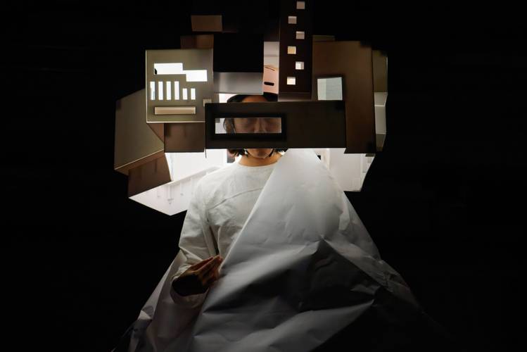 GPO Box No. 211 – Eine Person in weißem Gewand hält einen großen Papierbogen, Kopf von geometrischen Formen umgeben, auf schwarzem Hintergrund.