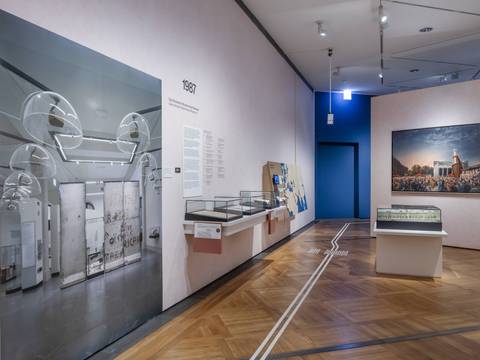  – Raumansicht der Ausstellung „Objekte. Geschichte. Geschichten. Blick in die Sammlung“. Auf der linken Seite ist ein etwa zwei Meter hohes Foto einer früheren Ausstellung des DHM zu sehen. Dahinter sind drei kleine Vitrinen sowie eine interaktive Landkarte zu erkennen. Rechts im Bild ist ebenfalls eine Vitrine im Raum zu erkennen, dahinter hängt ein Gemälde.