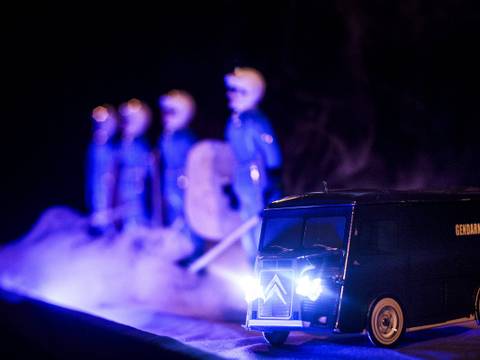 Blue – Spielzeuglieferwagen mit Aufschrift „Gendarmerie“ und leuchtenden Scheinwerfern. Im Hintergrund unscharfe Figuren in Uniformen mit Schwertern und Schilden.