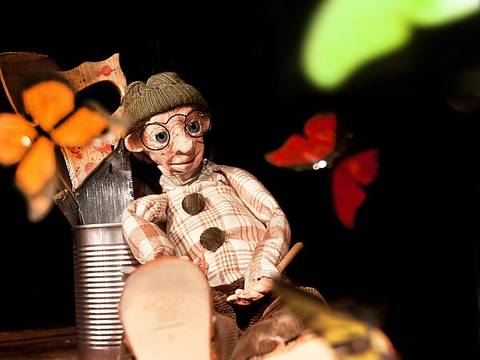  – Puppe mit Brille und Mütze sitzt auf Holzboden. Schmetterlinge fliegen umher. Konservendose mit bemaltem Holz dahinter.