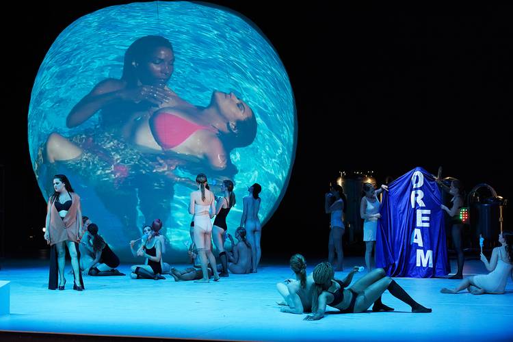 Die Entführung aus dem Serail – Personen in Bademode auf Bühne, große bläuliche Projektion von schwimmenden Personen, blauer Vorhang mit "DREAM" rechts, dunkler Hintergrund.