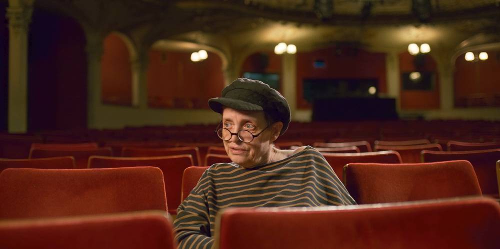 Helene Weigel - Revolutionärin im Rampenlicht – Person mit gestreiftem Pullover und Mütze sitzt in leerem Theatersaal mit rotem Samt. Hintergrund zeigt beleuchtete Bühne und verzierte Decke.