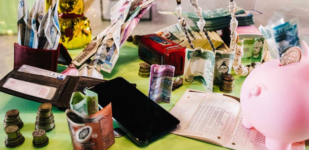 Buntes Stillleben mit Geldscheinen, Muenzen, rosa Sparschwein, Handy, Geldboerse, Kassenbuch und goldenem Winkekatze unter hell erleuchtetem Hintergrund.