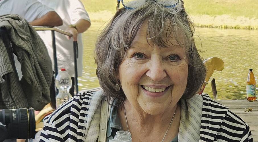 Die Lütte – Ein Abend für Angelika Mann – Ältere lächelnde Person mit grauem Haar und Brille auf dem Kopf, trägt eine gestreifte Jacke. Im Hintergrund ein Fluss und ein Tisch mit Flaschen.