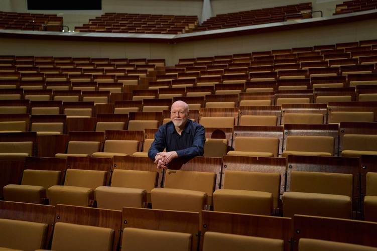 Brett Dean – Brett Dean sitzt allein in der Mitte eines großen, leeren Saals mit braunen Sitzreihen und blickt nach vorne, die Arme auf dem Sitz vor ihm gestützt.
