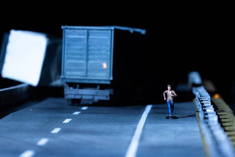 Transport: Cargo – Spielzeugfigur läuft auf Straße neben Spielzeuglaster, Kisten im Hintergrund, dunkle Umgebung.