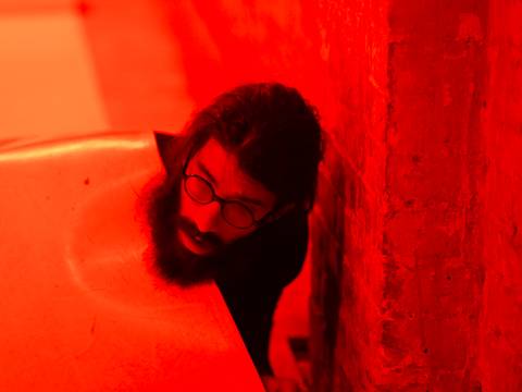  – Person mit Brille und Bart schaut hinter einer Wand hervor, in rotes Licht getaucht.
